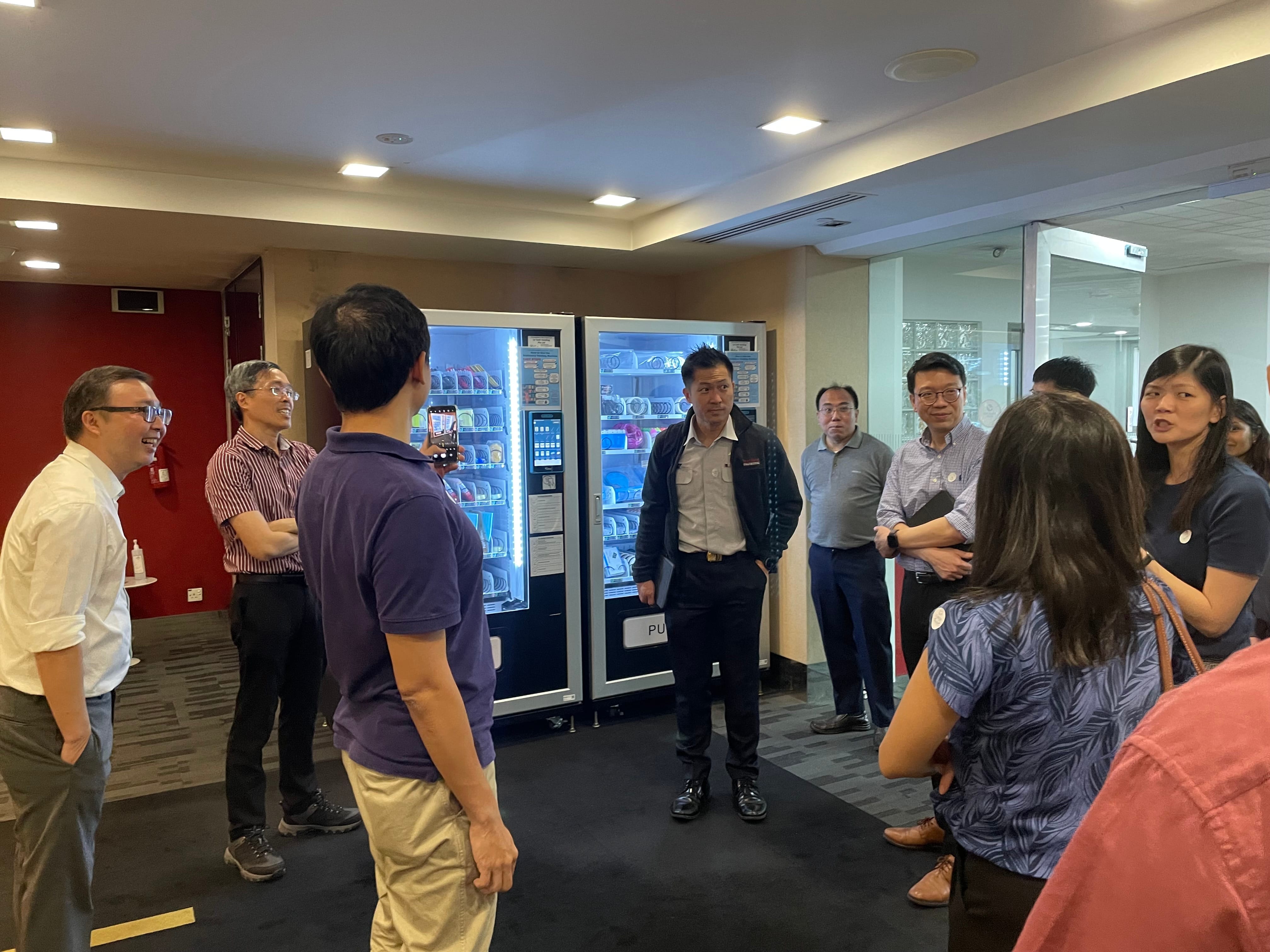 A group of people standing in a circle indoors, discussing near two lit vending machines stocked with items.